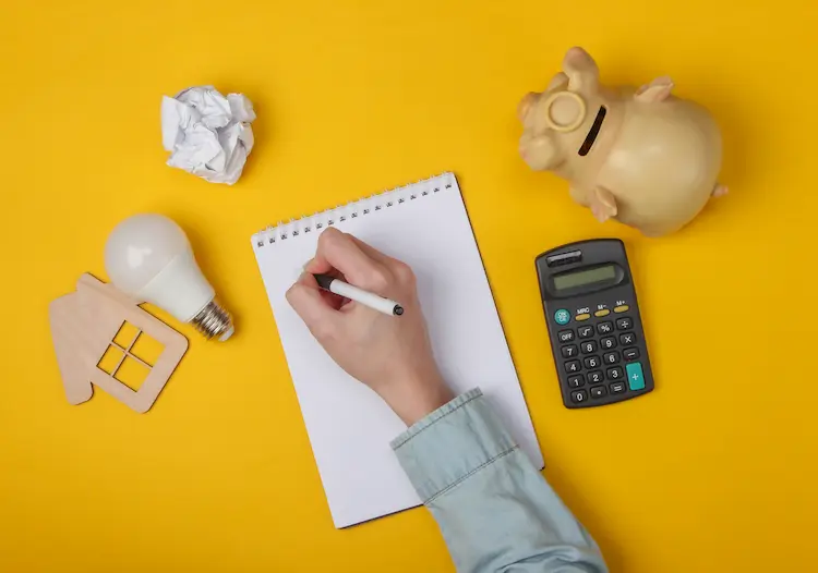 Dłoń pisząca w notatniku na żółtym tle, otoczona kalkulatorem, skarbonką-świnką, drewnianym domkiem i żarówką – wizualizacja procesu planowania finansowego.