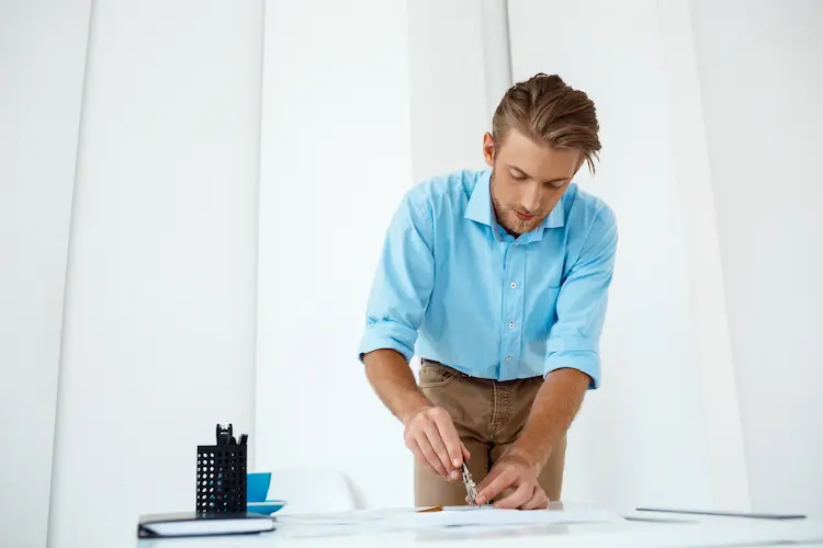 Mężczyzna w błękitnej koszuli używający cyrkla do precyzyjnej pracy nad dokumentem – wizualizacja procesu planowania sprzedaży w firmie.