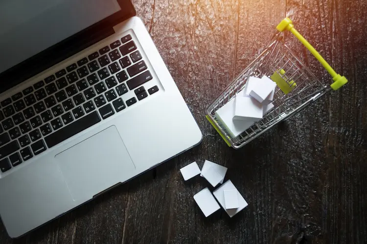 Laptop i miniaturowy koszyk zakupowy z białymi paczkami na drewnianym blacie – wizualizacja procesu skalowania e-commerce i wzrostu sprzedaży online.
