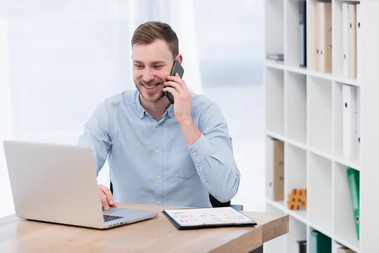 Uśmiechnięty mężczyzna w błękitnej koszuli rozmawia przez telefon przy laptopie i dokumentach – wizualizacja cech banku przyjaznego firmie.