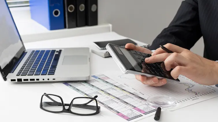 Właściciel firmy obliczający koszty logistyki e-commerce. Na pierwszym planie znajdują się okulary, laptop. kalkulator i arkusze kalkulacyjne.