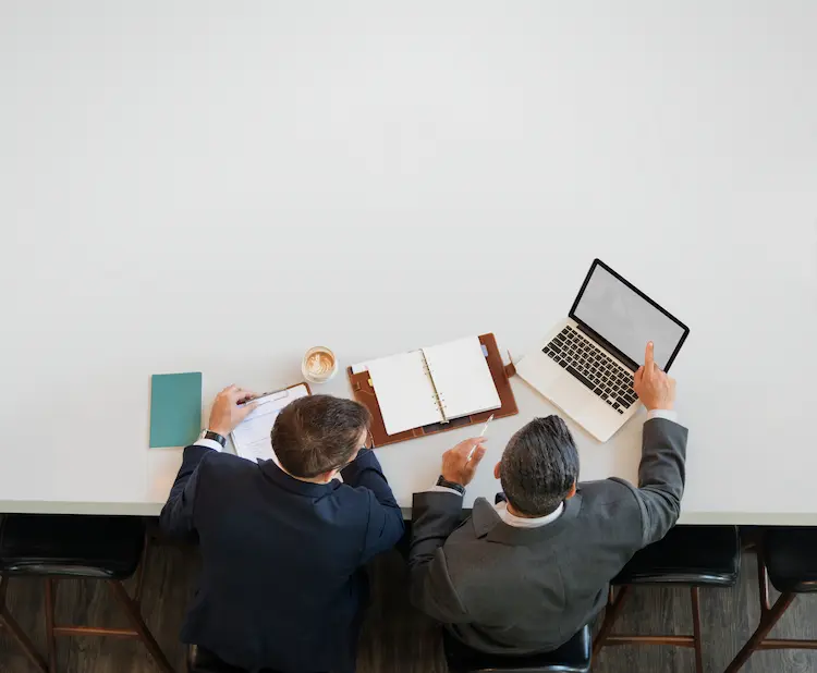 Dwaj biznesmeni przy białym biurku analizujący na laptopie korzyści z założenia fundacji rodzinnej – widok z góry.