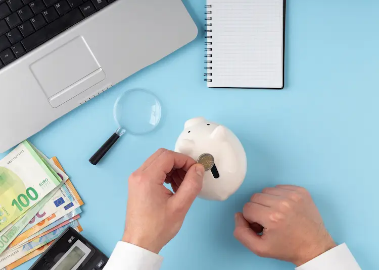 Widok z góry na biurko z laptopem, kalkulatorem i banknotami euro, dłonie wrzucające monetę do białej świnki skarbonki obok lupy i notesu.