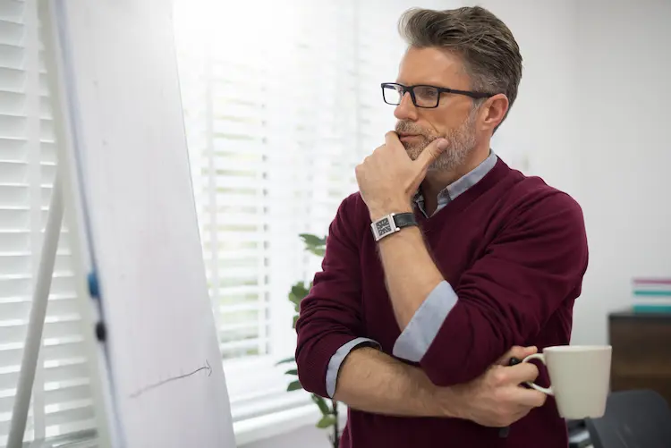 Dojrzały mężczyzna w okularach i bordowym swetrze patrzy z namysłem na białą tablicę z wykresem, trzymając kubek kawy w jasnym biurze.