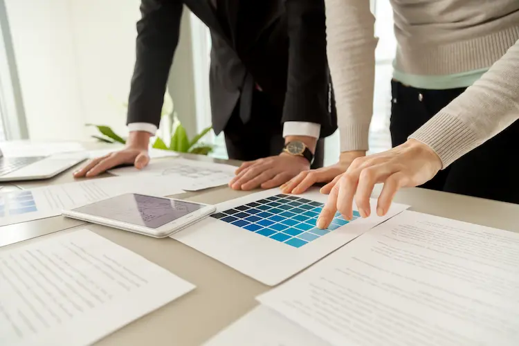 Dłonie dwóch osób pracujących przy biurku nad dokumentami; jedna osoba wskazuje palcem na niebieską siatkę kolorów lub wykres na arkuszu papieru obok tabletu.