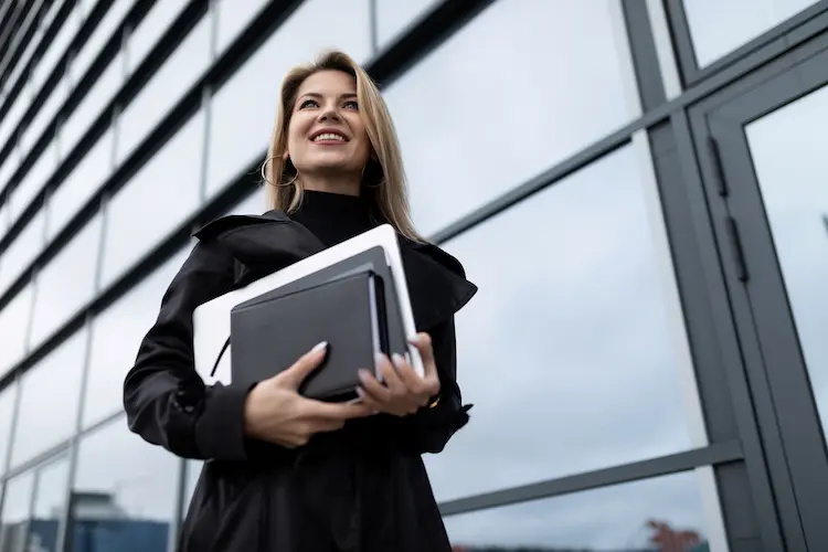 Uśmiechnięta kobieta sukcesu przed szklanym biurowcem, trzymająca laptopa i notatnik – wizualizacja korzyści, jakie dają studia MBA w Polsce.